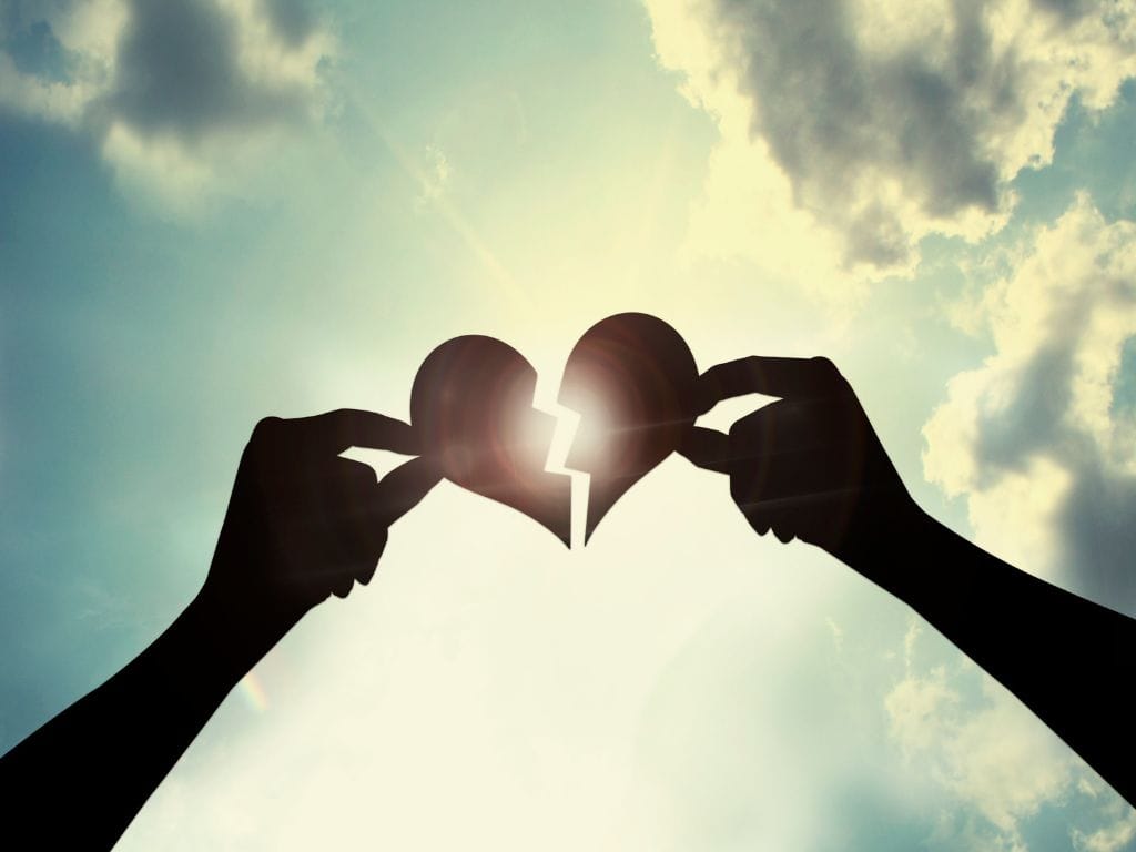 Broken heart with each half held by a hand and being put together with sun and sky in the background
